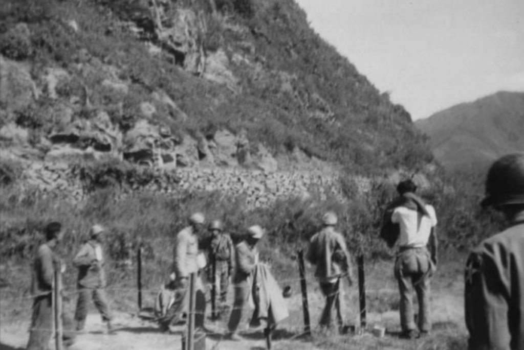 A black and white photograph shows a few Korean prisoners walking with American soldiers. They appear to be inside a barbed wire area in front of a mountain.