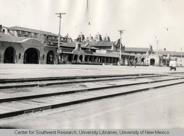 A black and white photograph of the outside of a massive hotel complex. In the front are several railroad tracks.