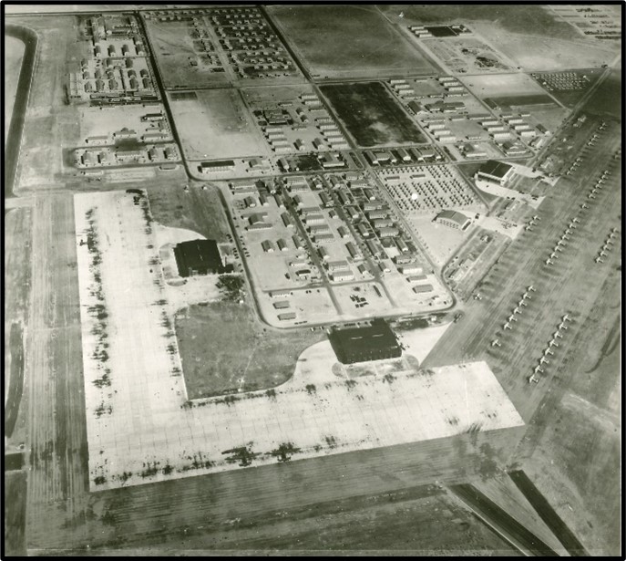 A black and white aerial image of an airbase. To the front and left is an airstrip. To the right are many columns of airplanes in line.
