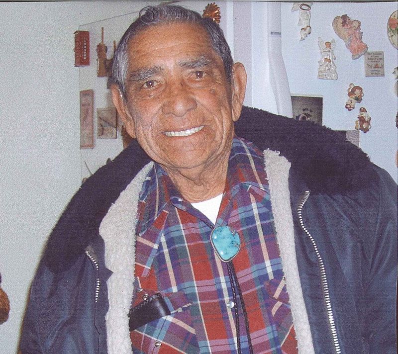 A color photograph showing an older man from the chest up. He is wearing a plaid shirt covered by a blue jacket. He is smiling at the camera.