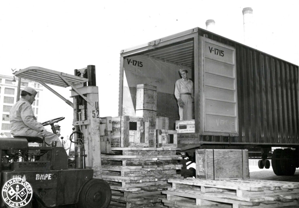 A black and white photograph of two white soldiers. One is running a forklift, putting materials inside a box truck. The other is inside the box truck. 