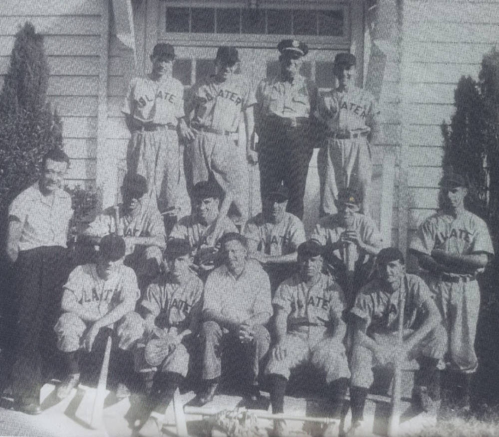 A black and white photograph showing a baseball team of white men in baseball uniforms that say ‘Slater’ across the front. They are posing in front of a house. 