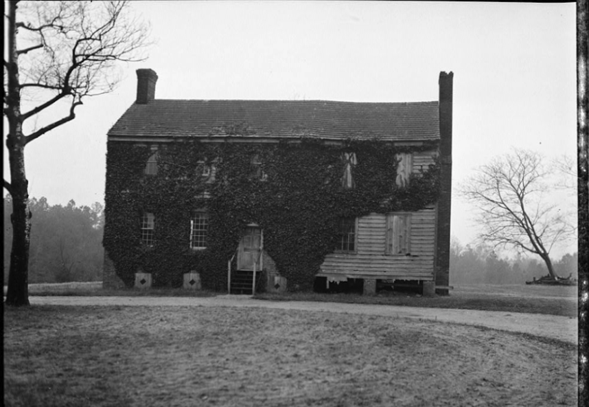 A black and white photo of a run-down, two-story, clapboard home mostly covered in vines. There is a circular driveway in the front and a couple of trees on the property. 