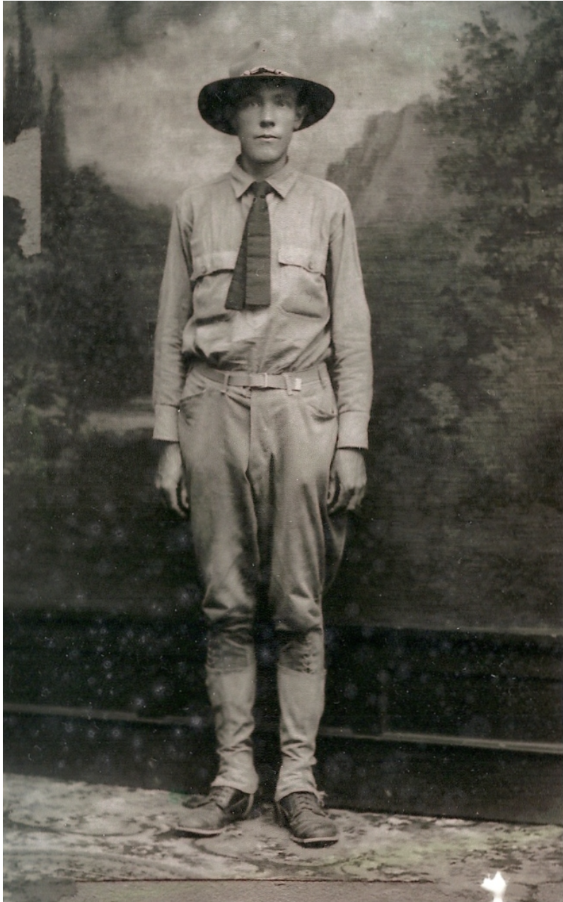 A black and white photograph of a White young man in uniform standing in front of a scenic painted backdrop.