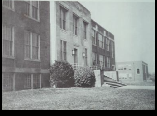 A black and white photo of the side view of a three-story brick schoolhouse. There are two large bushes next to the stairs and a front lawn in front of the school. 