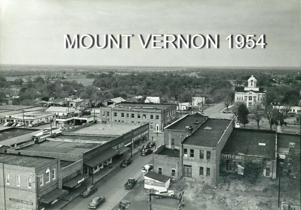 A black and white aerial image of a city's main street, showing several buildings and vehicles. 