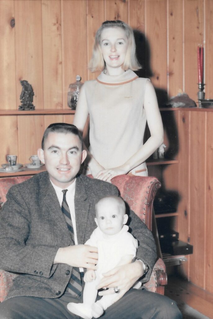 A color image of a White couple - man and woman with a baby. The woman has shoulder-length blonde hair and stands behind the chair that the man is sitting in. They are both in dressy clothes. The baby sits on the man's lap in the chair. The image is taken inside the house.