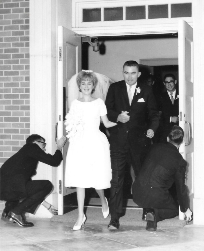 A black and white image of a White couple leaving the wedding chapel. The man is in a black suit. The woman is in a knee-length, white wedding gown. Two men are kneeling to prop the doors open on either side of them.