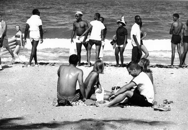A black and white image of several Black men in swimsuits at the beach, while White beachgoers look at them. 