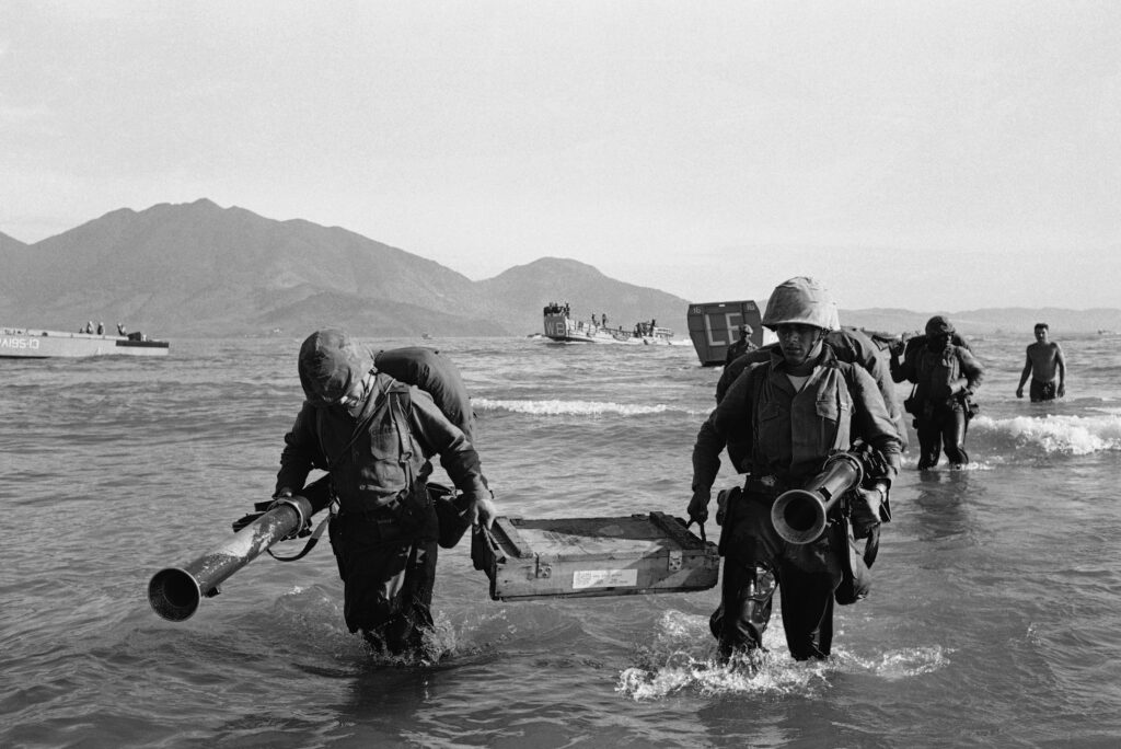 A black and white photograph of a handful of Marines wading through knee-high water carying supplies from a boat to the shore. In the distance are other boats and a mountain range.