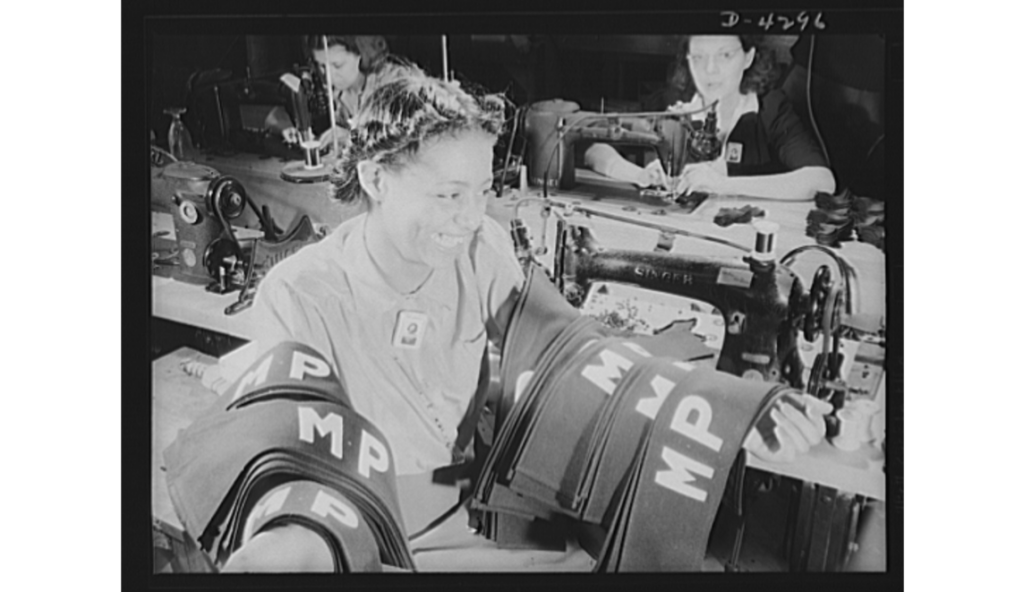 A black and white image of a Black young female sitting at a sewing machine with her arms covered in “MP” bands.