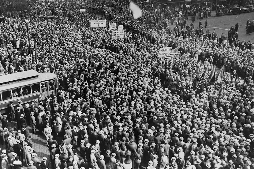 A black and white aerial image of thousands gathered in an outdoor protest with signs. 