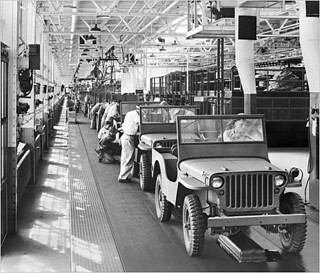 A black and white image of an assembly line of Jeeps being worked on in a large factory. 