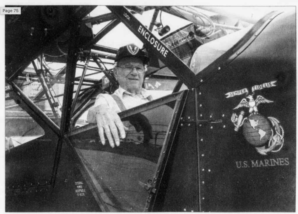 A black and white image of an older white man sitting in the cockpit of an older airplane. The USMC logo is painted on the side of the plane.