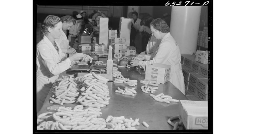 A black and white image of several white women in white lab coats and hairnets standing around a metal table piled with sausage they are weighing and packaging.