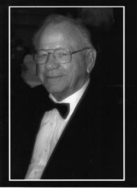 A black and white image of a older White man. He is wearing a black suit and bowtie. He appears to be seated and the photo is from the chest up.