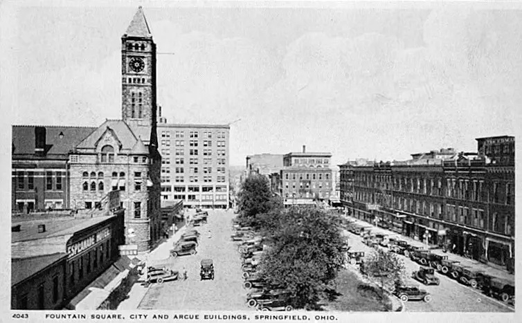 A black and white aerial shot of a city street with many Model-A type vehicles parked on the street. There are several multi-story, brick buildings. 