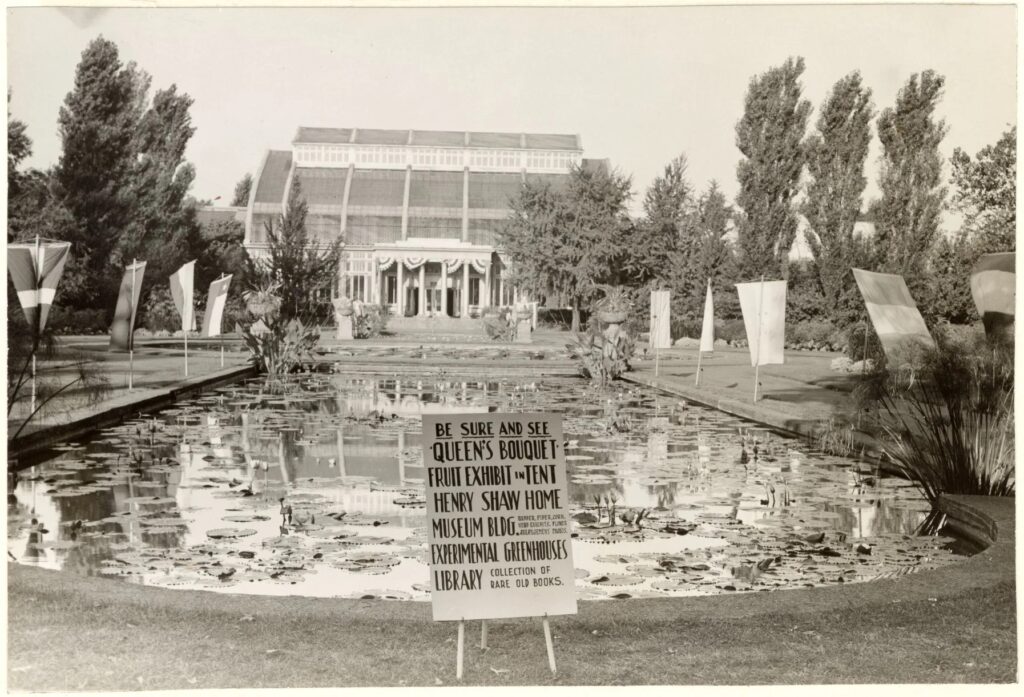 A black and white image of a three-storm building with a columned entrance in the backyoung. It is decorated with patriotic bunting. In front is a decorative pool filled will lily pads and flags of our allies around the sides. In from is a sign that reads, “Be sure to see Queen’s Bouquet Fruit Exhibit in Tent Henry Shaw Home Museum BLDG. Experimental Greenhouses Library.”