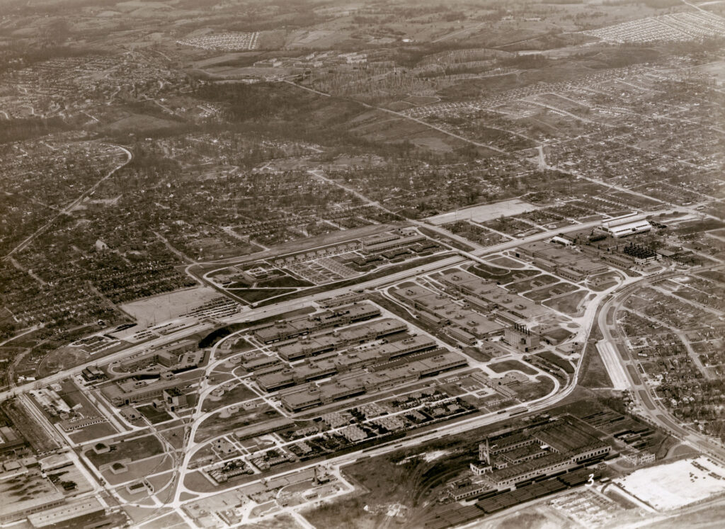 A sepia-toned aerial image showing multiple factory buildings in a row on the outskirts of the city and suburban area. 