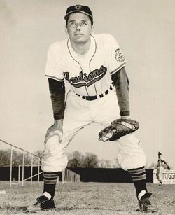 A black and white image of a White man in a baseball uniform. He is bent over with a mit on one hand.