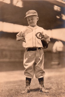 A sepia-toned photo of a little boy standing on a baseball field in a Chicago Cubs uniform. 