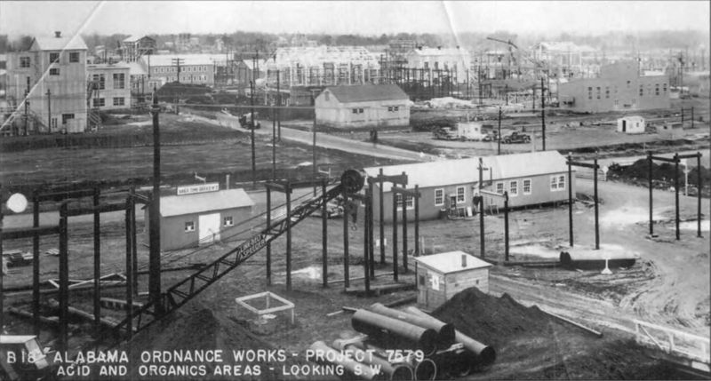 A black and white image showing a crane in the foreground. There are several framed structures going up on the construction site. 