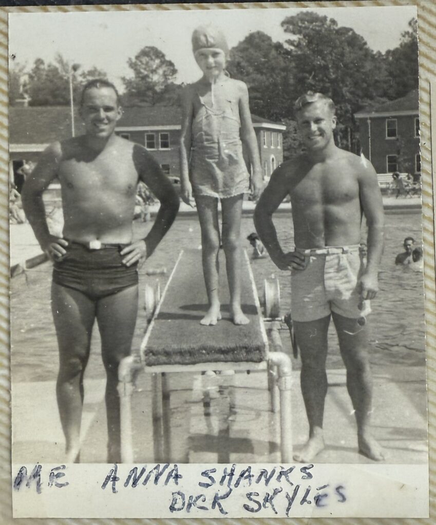 A black and white image of two white men standing in bathing suits alongside a white girl on a diving board. There are other people swimming in the pool behind them. 