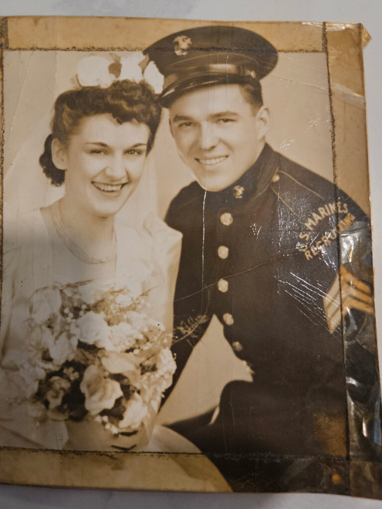A sepia-toned image of a seated bride and groom. The bride is in a white dress with a floral veil. She is holding a large bouquet of flowers. The groom is in his uniform. 
