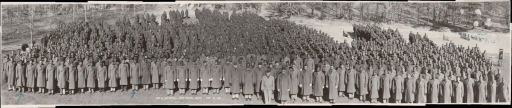 A black and white elevated image of several hundred soldiers in long coats standing in a field in formation. 