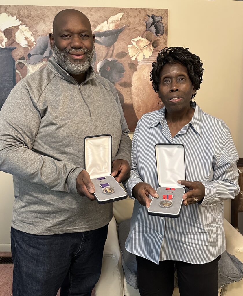 A color photo of two older Black people (a man and a woman) each holding a box with a medal inside.