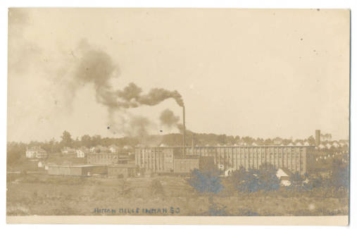 A sepia photograph of mill buildings taken from a distance. The smoke stack is pumping out lots of smoke.