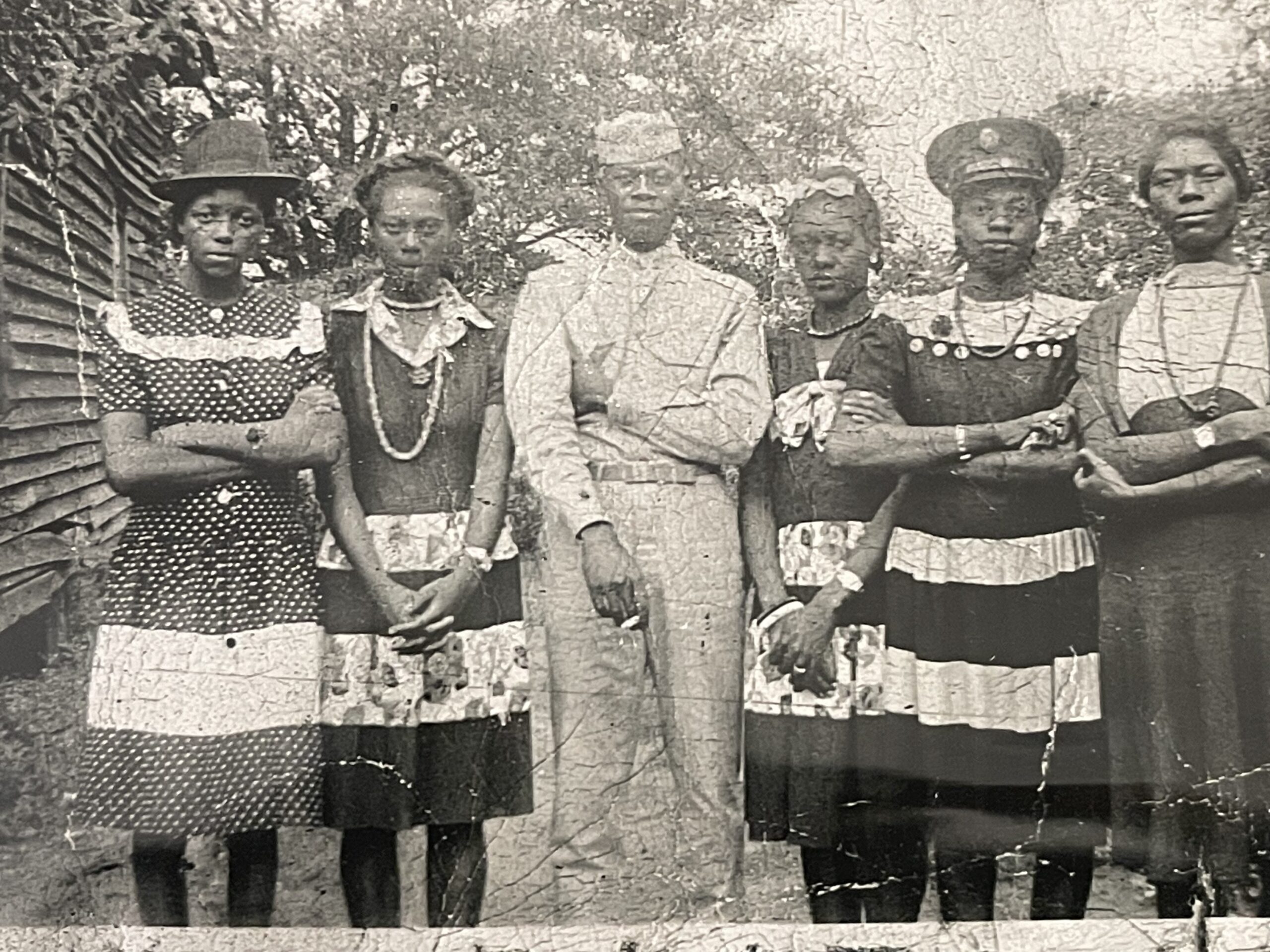 A black and white image of five young Black women standing around one young Black man in uniform. The women are all in dresses. They are standing outside a house. 