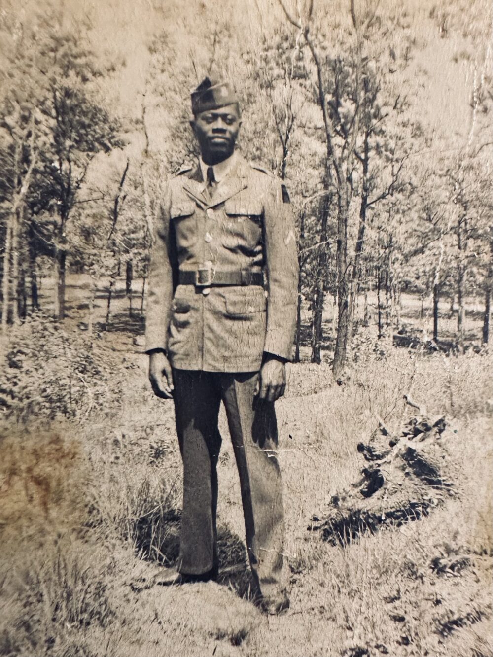 A sepia photograph of a young Black man in uniform standing in a field surrounded by trees.