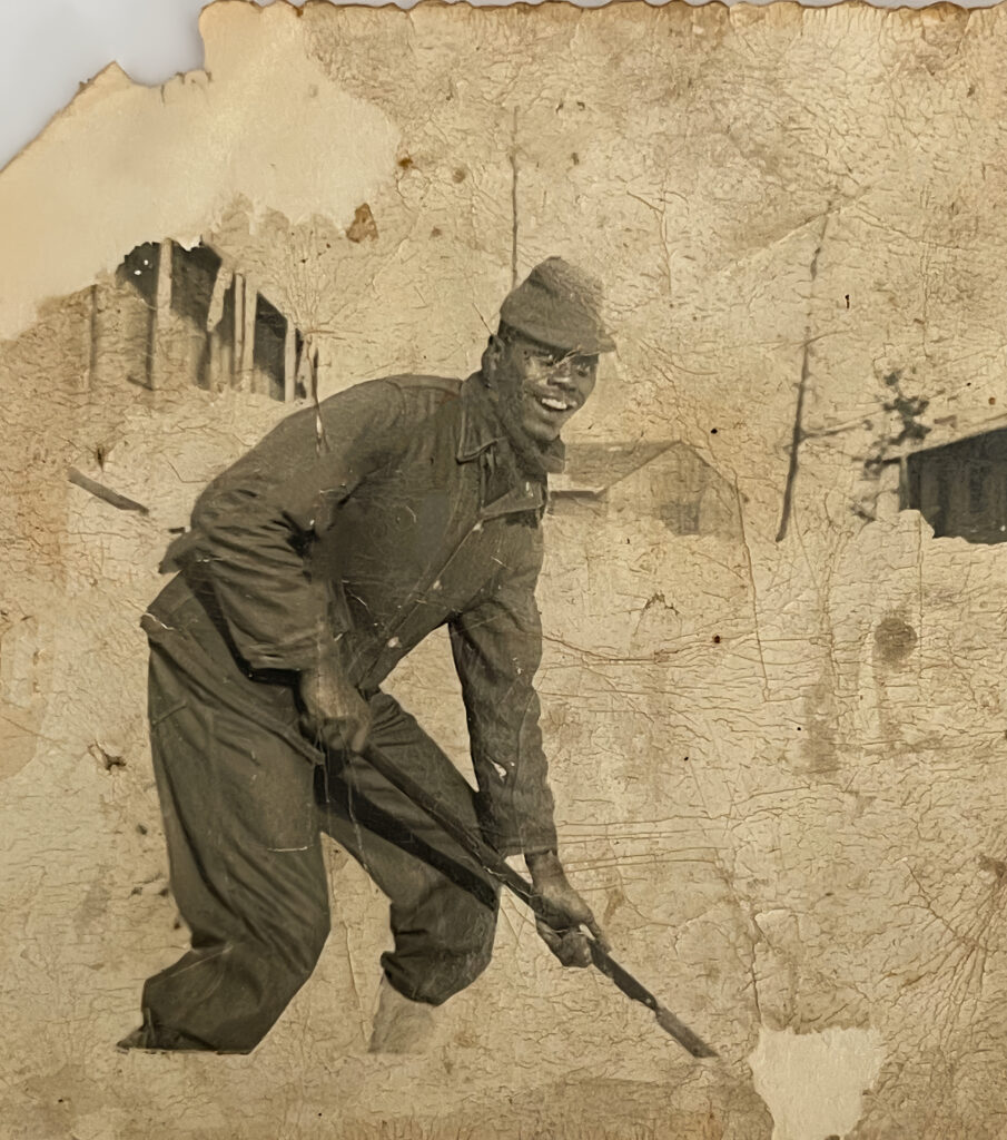 A sepia image of a young Black man in uniform bending over with a shovel in his hands. The background of the image has been faded. 