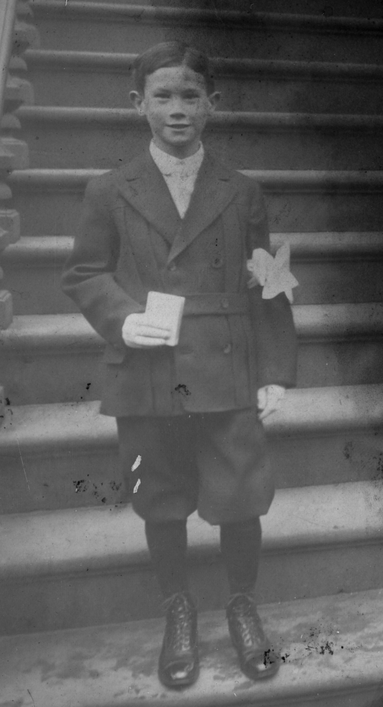 A black and white photo of a white boy standing at the landing of stairs in short, wool pants with knee-high socks, and a wool dress jacket. He is holding a bible in his right hand and there is a white decorative flower on the left arm.