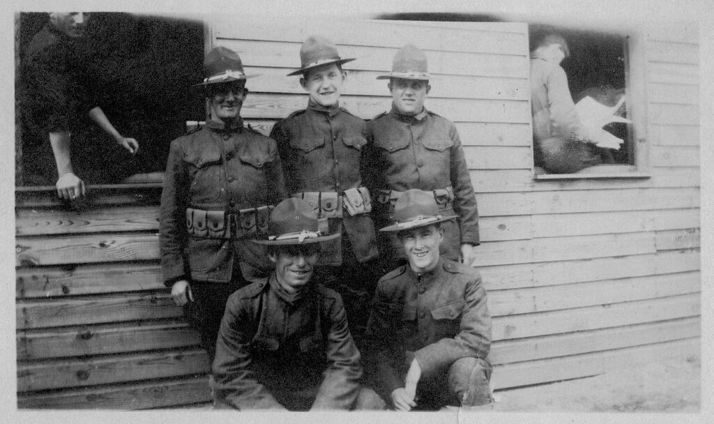 A black and white image of give white men in uniform standing outside a wooden house. On either side there are two windows with one man sitting in each.