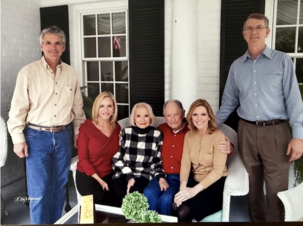 A color photo. On a white wicker bench in the center sits an elderly Ike and Kathryn Rigell. On either side are their two daughters. Standing on either side of the bench are their two sons.