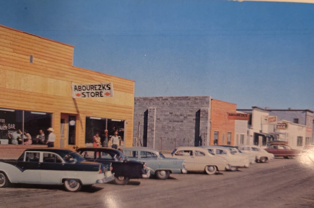 A color photo of a main street of stores with cars parked on the street.