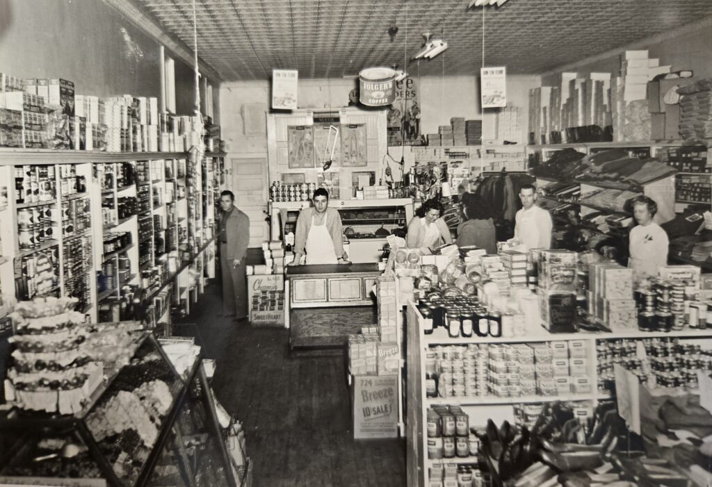 A black and white image of a grocery store with several customers and workers inside.