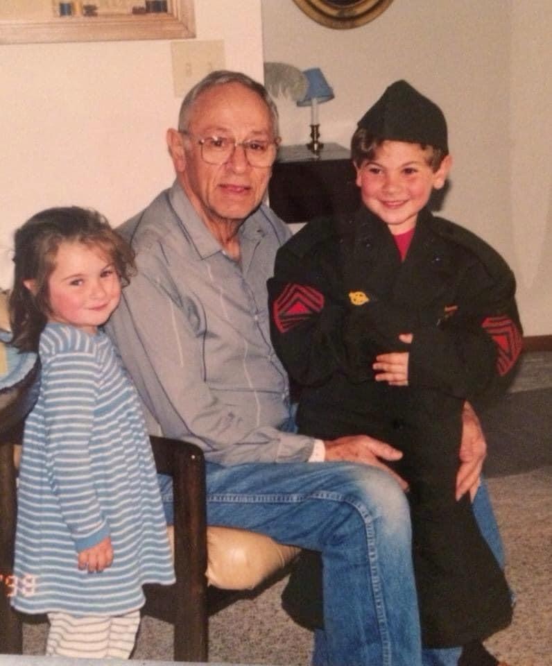 A color photo of an elderly White man sitting in a chair in between two young kids, a girl and a boy.