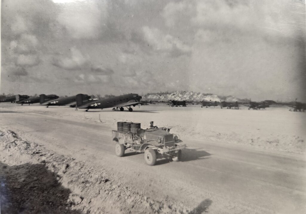 A black and white photo of a military vehicle transporting vehicles on an airfield. In the background are several airplanes.