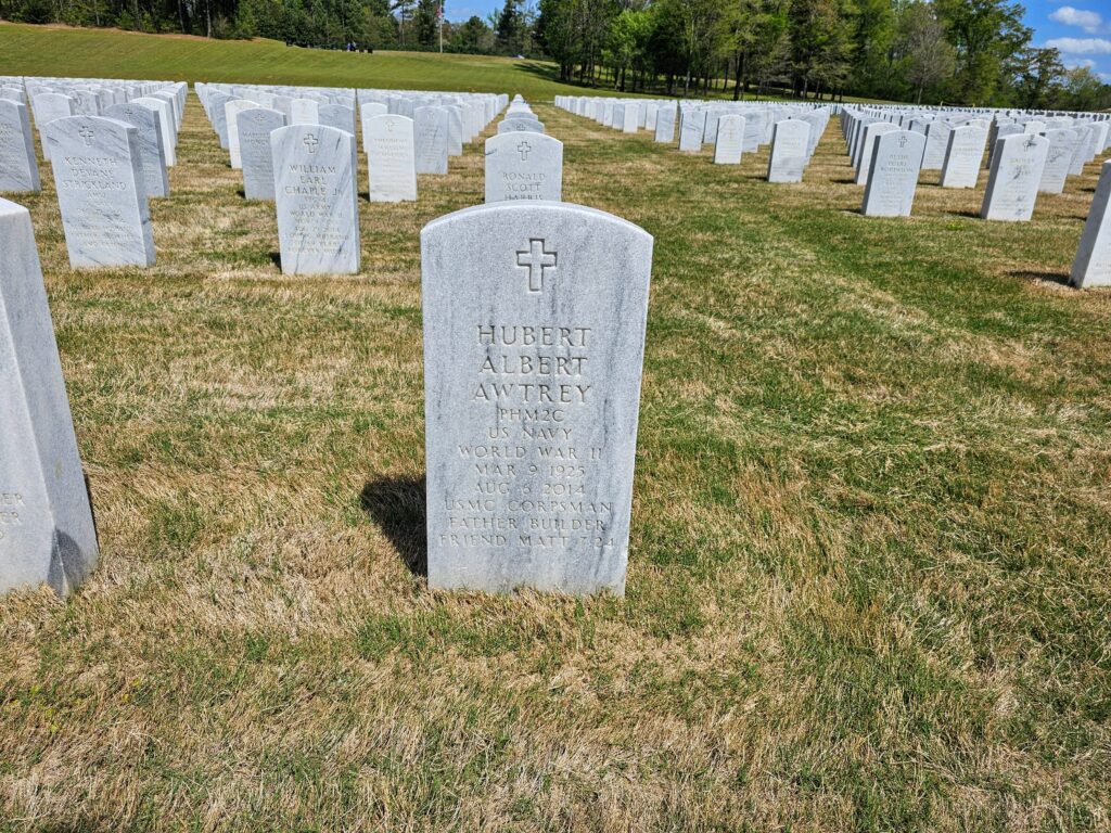 An arched gray marble headstone engraved with a cross and the words "Hubert Albert Awtrey PHM2C US Navy World War II Mar 9 1925 Aug 5 2014 USMC Corpsman Father Builder Friend Matt 7-24"