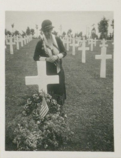 A black and white image of a woman in a dress and hat standing next to a cross headstone that is covered with flowers and an American flag.