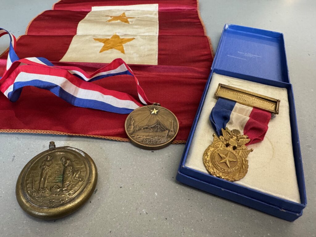 A red flag with two gold stars, a bronze medal with the Statue of Liberty and Eiffel Tower as well as a ship and the year ‘1932’, A gold medal on red, white, and blue ribbon with Emma’s name engraved is in a box. One other medal with two figures on it.
