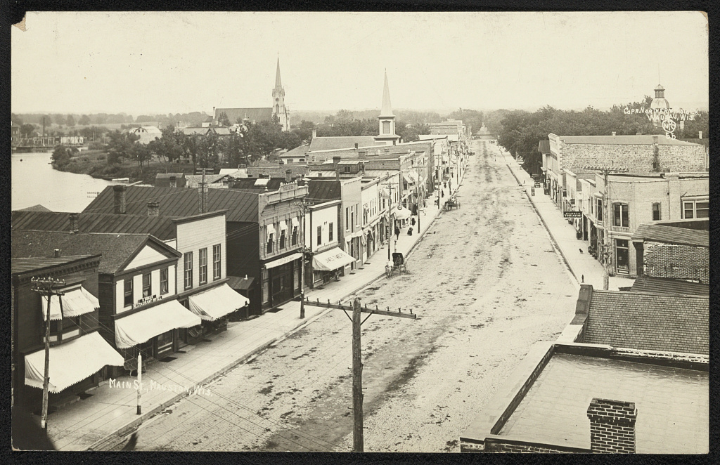 A black and white aerial photo of a city street. Features several two story buildings. Several church steeples can be seen in the background.