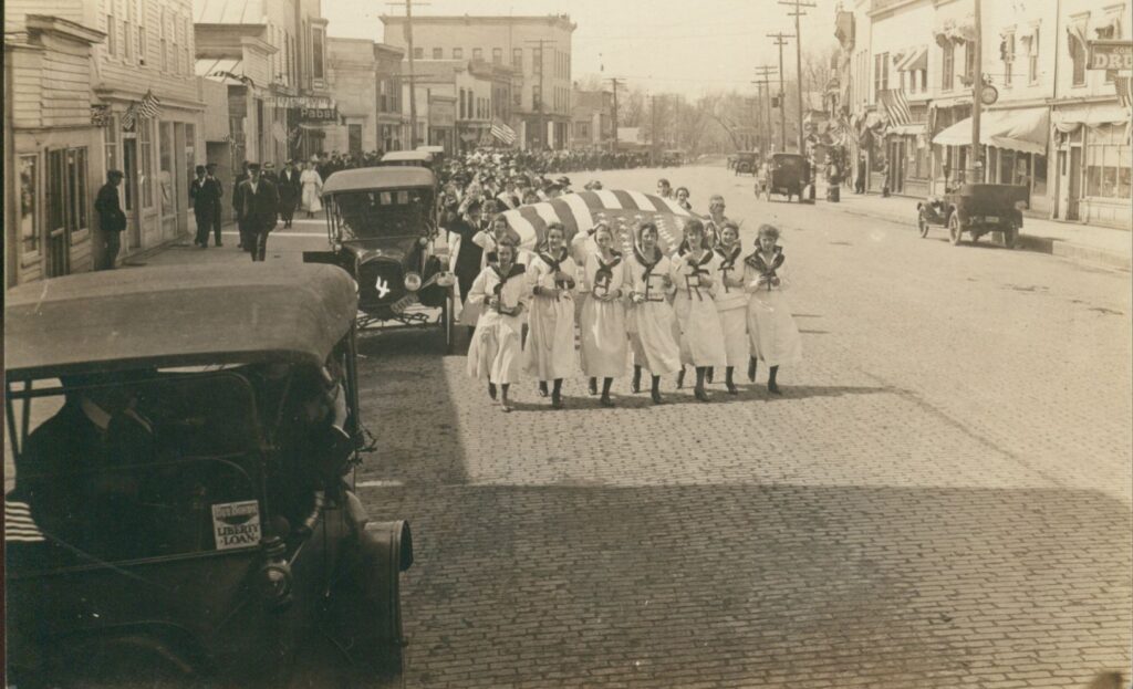 A black and white image of several dozen people in a parade down a brick city street. The girls in the front of the parade are holding a large American flag. They are dressed all in White.