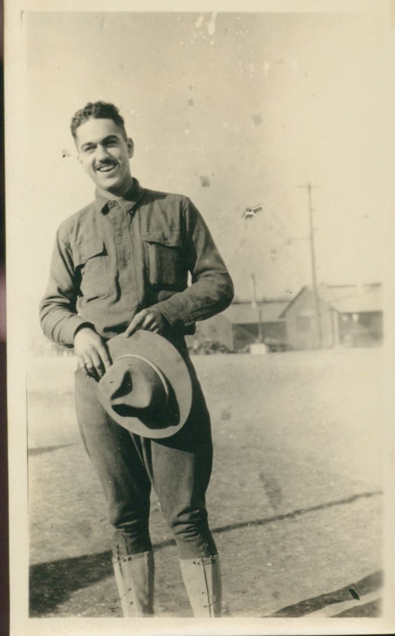 A black and white photo of a young White man standing in the street outside some training barracks. He is uniform with his hat in his hands and is smiling.