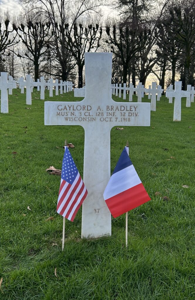A white marble cross is engraved with “Gaylord A. Bradley Mus’n 3 Cl. 128 Inf. 32 DIV Wisconsin Oct. 7 1918.” Next to the cross in the ground are a small American and French flag.