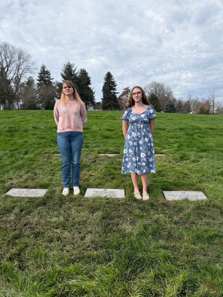 Two young White female students stand in a cemetery.