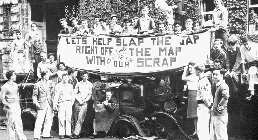 A black and white image of several young people standing around cars with the banner on top that reads, "Let's Help Slap the Jap Right Off the Map With Our Scrap."